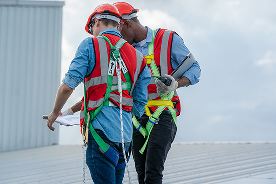 Two roofing workers in PPE and safety harnesses stand on a metal roof while they look at a document one holds.