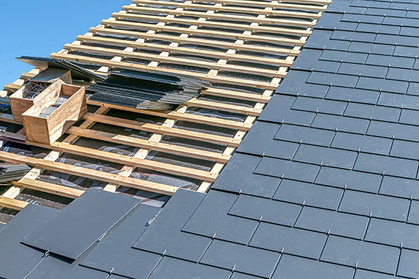 A slate roof mid-installation, with a stack of slate tiles on top of bare wood to the side of the completed roof section.